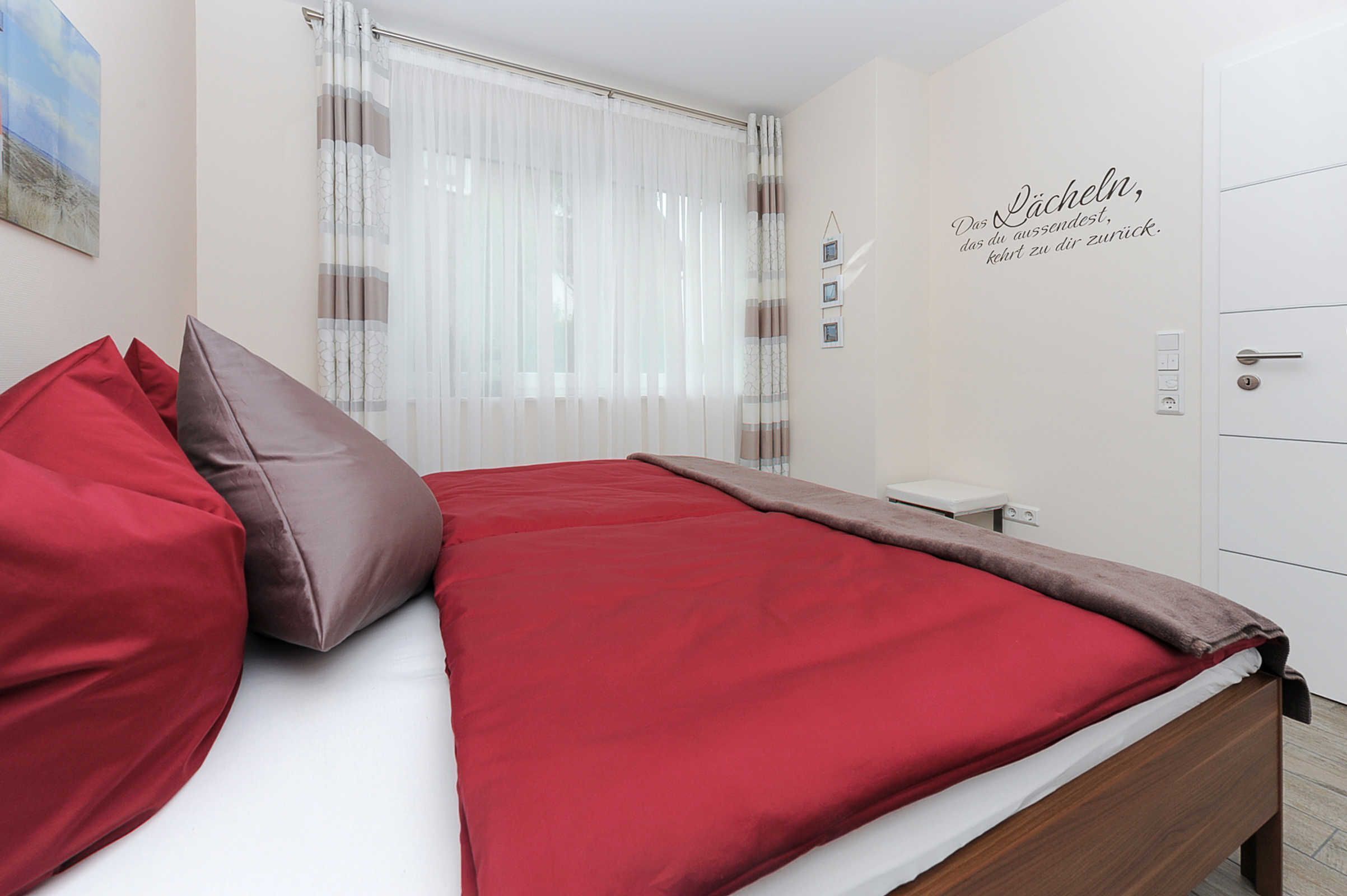 A neatly made bed with red and brown bedding, a bedside table with a lamp, and a wall with a decorative sign.