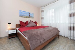 Ein Schlafzimmer mit einem hölzernen Bett, Bettwäsche in Rot- und Brauntönen, einem Nachttisch mit Lampe, einem Wandbild und leichten Vorhängen.