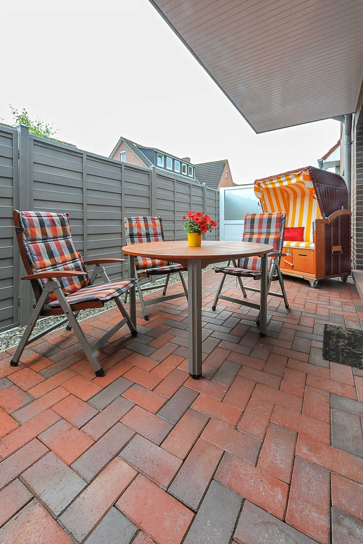 Eine Terrasse im Freien mit rotem Backsteinboden, einem runden Tisch und zwei Stühlen mit karierten Sitzkissen.