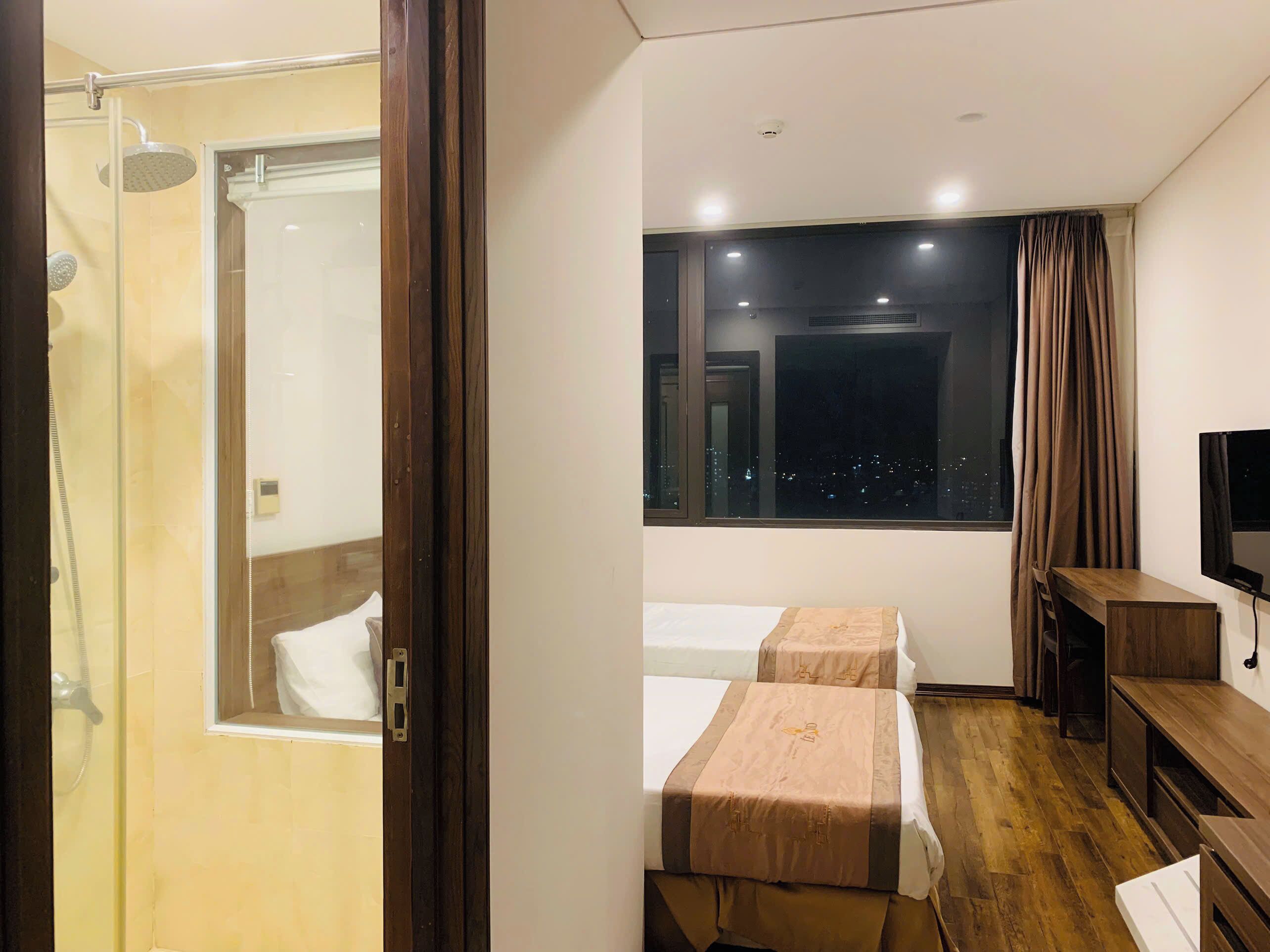 Ein Hotelzimmer mit Dusche, zwei Betten, Fernseher und Fenster mit Blick auf die beleuchtete Stadt.