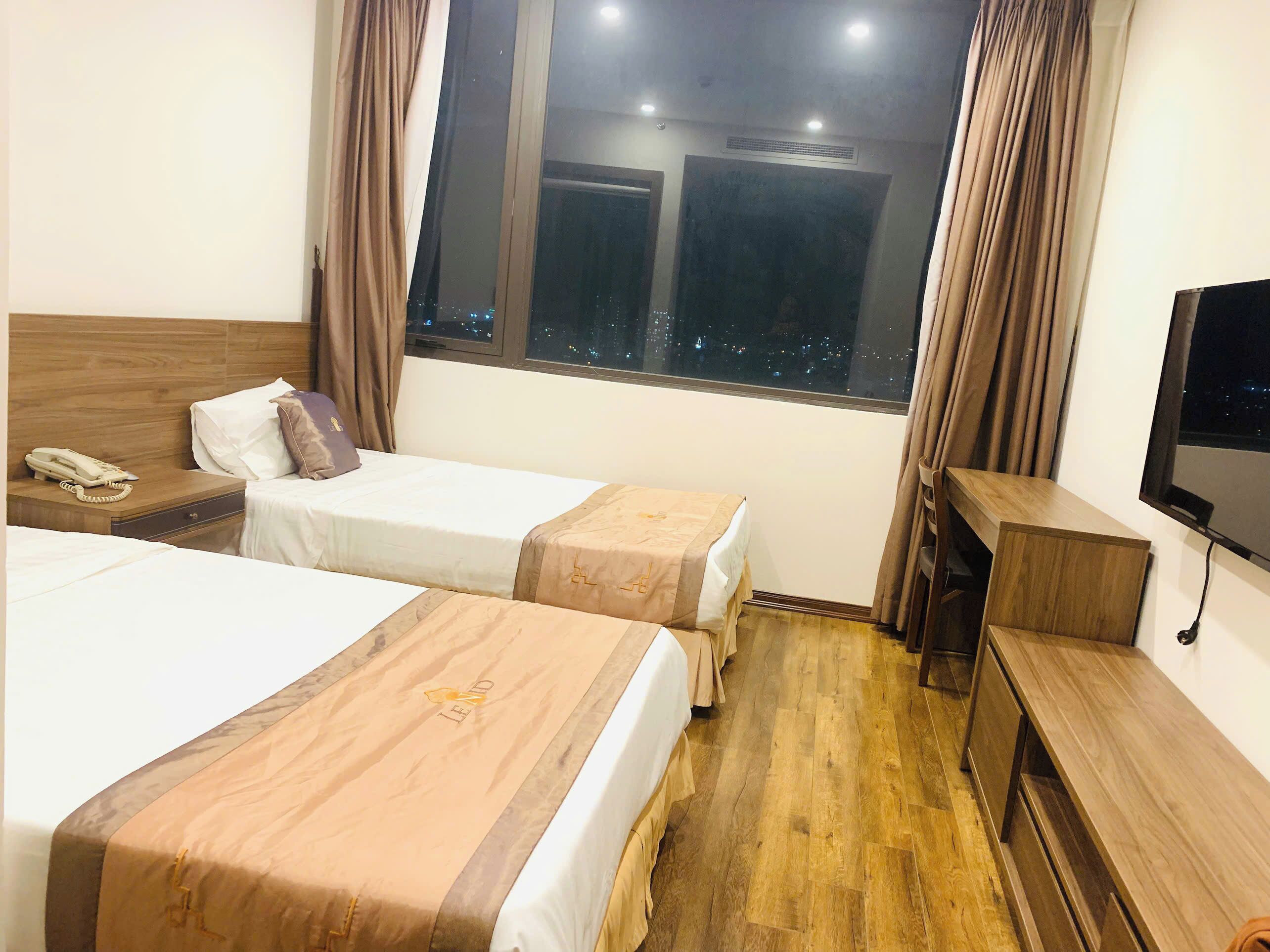 Ein Hotelzimmer mit zwei Einzelbetten, einem Nachttisch aus Holz mit Telefon, einem Holztisch mit Fernseher und einem großen Fenster mit Blick auf die beleuchtete Stadt.