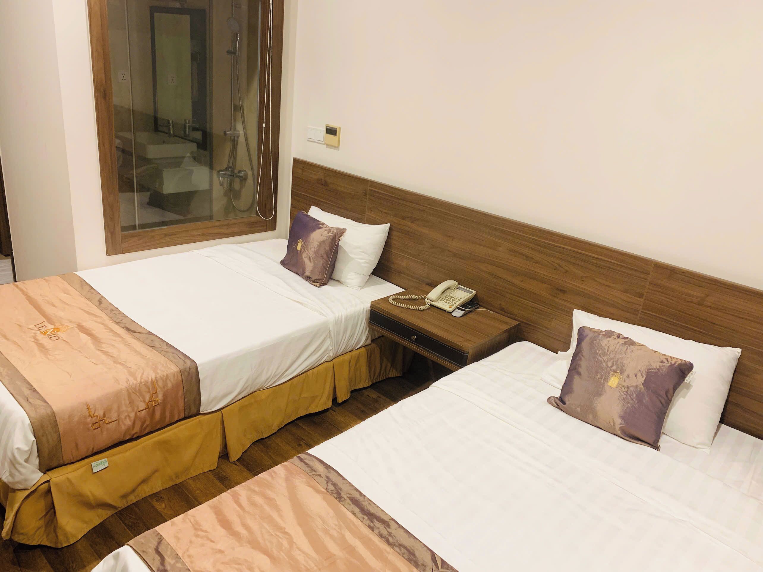 A hotel room with two beds, each with a wooden headboard, a bedside table with a telephone, and a window with a view of a bathroom.