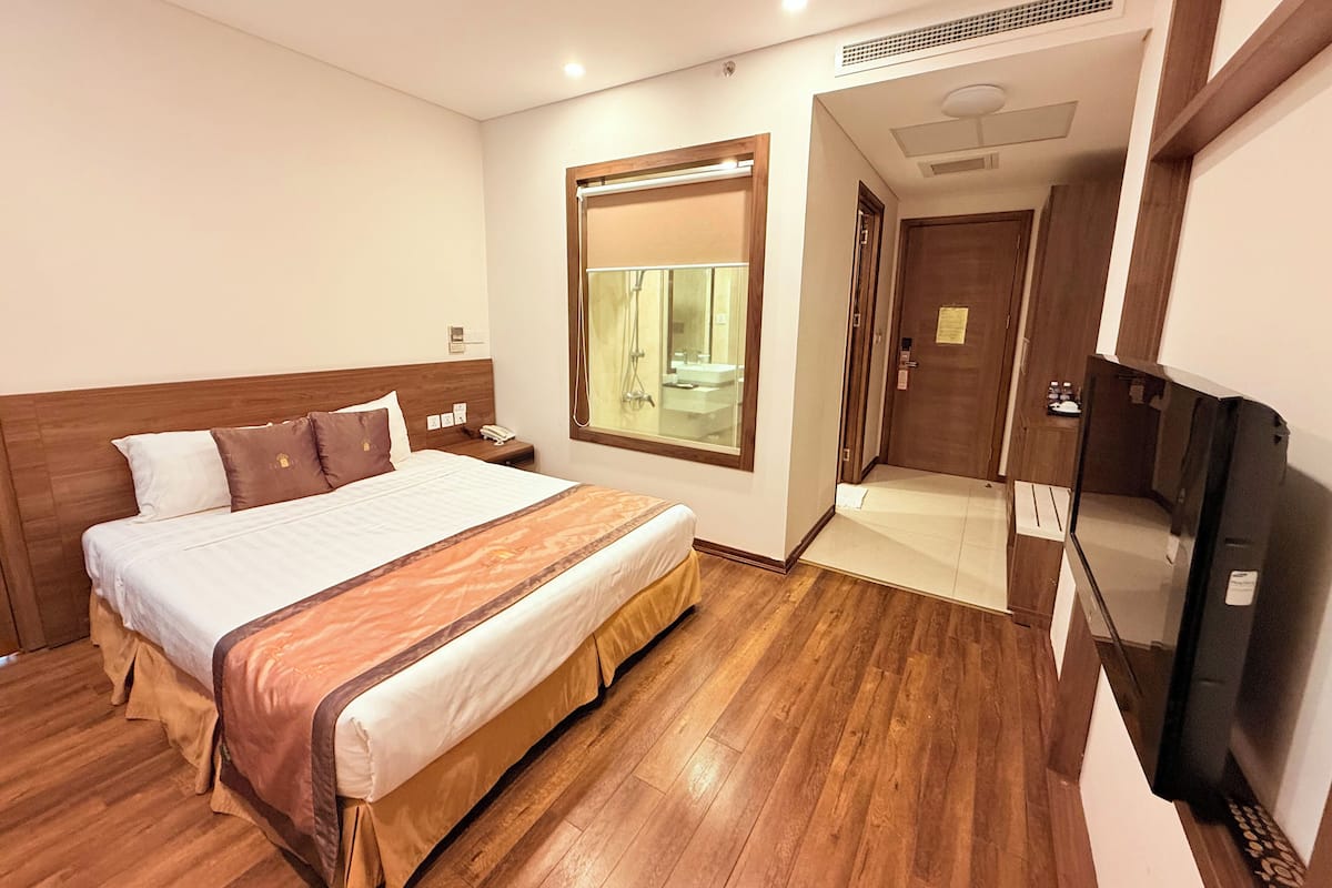 Een hotelkamer met een houten vloer, een groot bed met hoofdbord, een televisie, een raam met uitzicht op een badkamer en een deur naar een andere kamer.