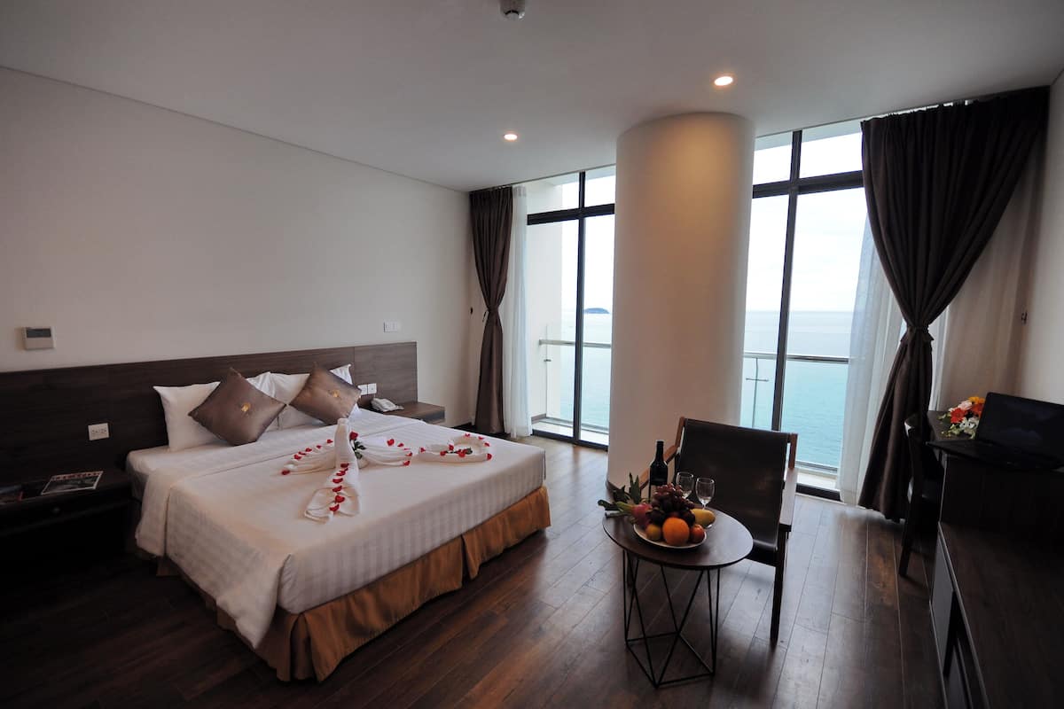 Een hotelkamer met een groot bed, een tafeltje met fruit en wijn, en uitzicht op de zee door grote ramen.