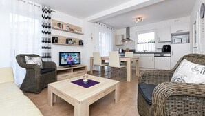 Ein modernes Wohnzimmer mit einer Couch, einem Couchtisch, einem Fernseher und einem Essbereich mit Stühlen sowie einer Küche im Hintergrund.
