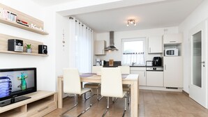 Eine moderne Küche mit weißen Schränken, einer Mikrowelle und einer Kaffeemaschine. Ein Esstisch mit Stühlen und ein Fernseher auf einem Holzgestell sind ebenfalls vorhanden.