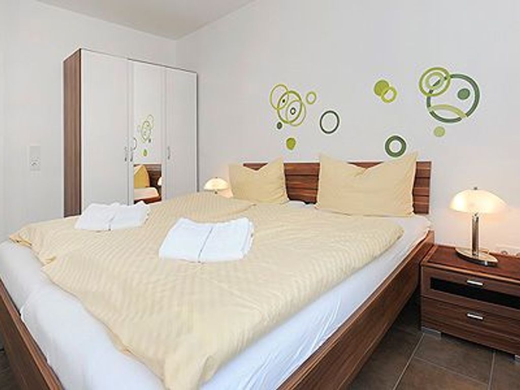Ein ordentlich bezogenes Bett mit weißer Decke und Kissen, ein Nachttisch mit Lampe sowie ein Kleiderschrank mit Spiegel.
