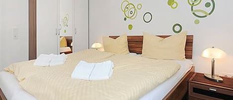 Una cama bien hecha con edredón blanco y almohadas, una mesita de noche de madera con una lámpara y un armario con espejo.