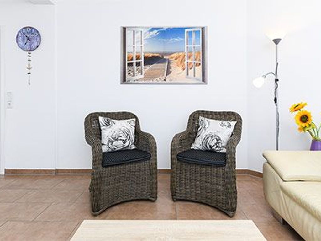 Una habitación con dos sillones de mimbre con cojines negros, un sofá beige, un reloj de pared, un cuadro enmarcado de una playa y una lámpara de pie con un jarrón de girasoles.