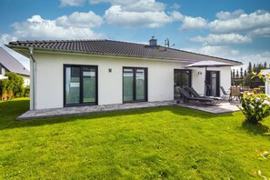 A modern two-story house with a flat roof, large windows, and a patio with outdoor furniture.