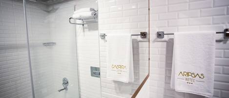 Un baño moderno con ducha envuelta en vidrio, inodoro y lavamanos. Hay toallas colgadas en un perchero y una toalla con el texto "ARIPSA HOTEL".
