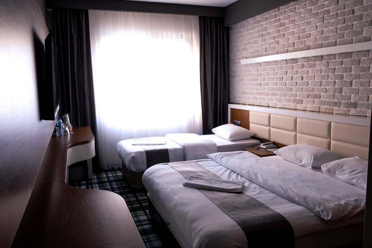 Een hotelkamer met twee bedden, een bakstenen muur, een raam met gordijnen en een bureau met een lamp en waterflessen.