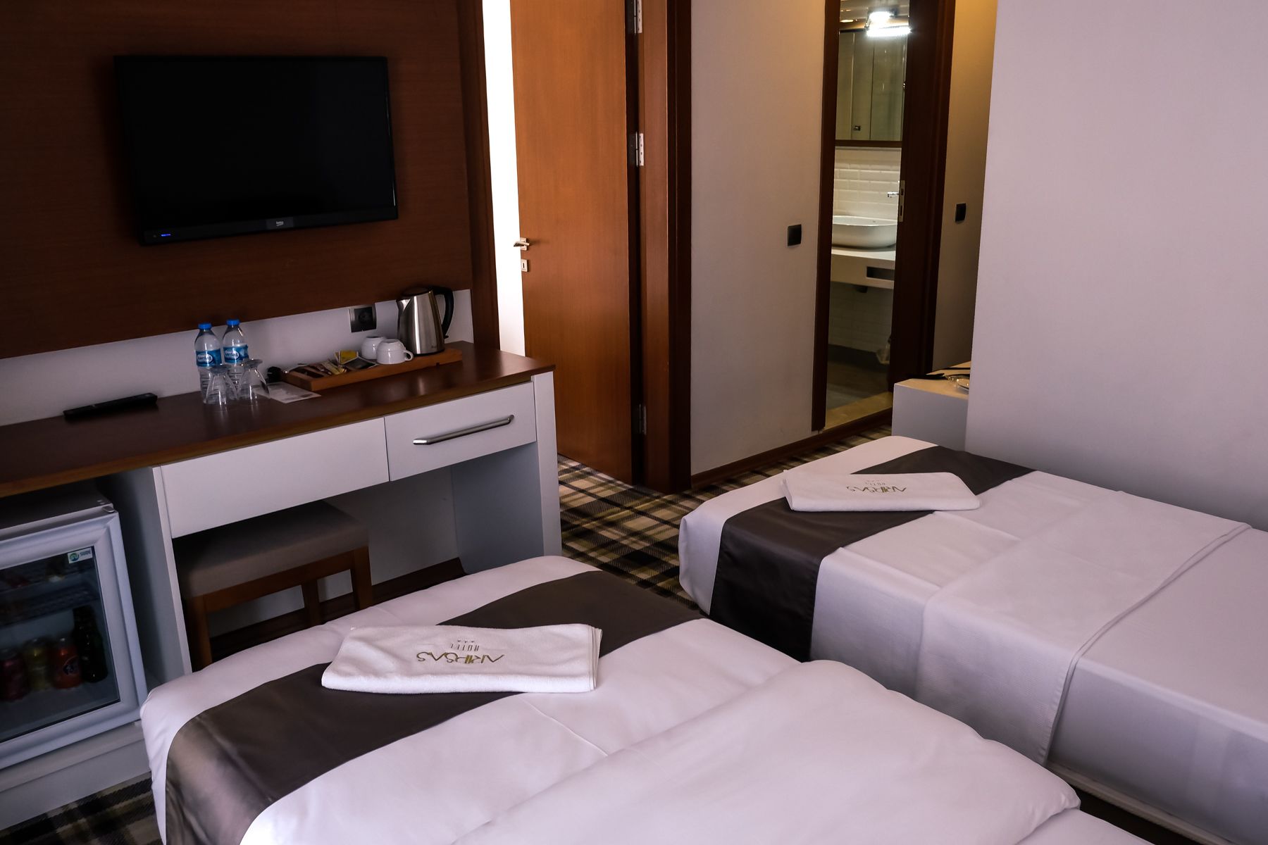 Habitació d'hotel amb televisió, escriptori amb bullidora, dos llits amb tovalloles i nevera.