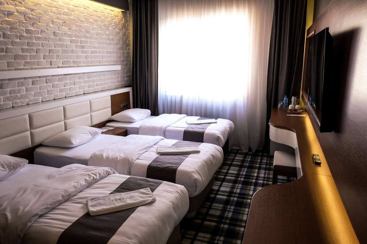 Hotelkamer met drie bedden, een bakstenen muur, een groot raam met gordijnen, een houten bureau en een televisie.