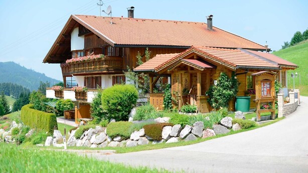 Un chalet in legno con tetto spiovente, un balcone con fioriere e un vialetto in pietra che conduce all'ingresso.