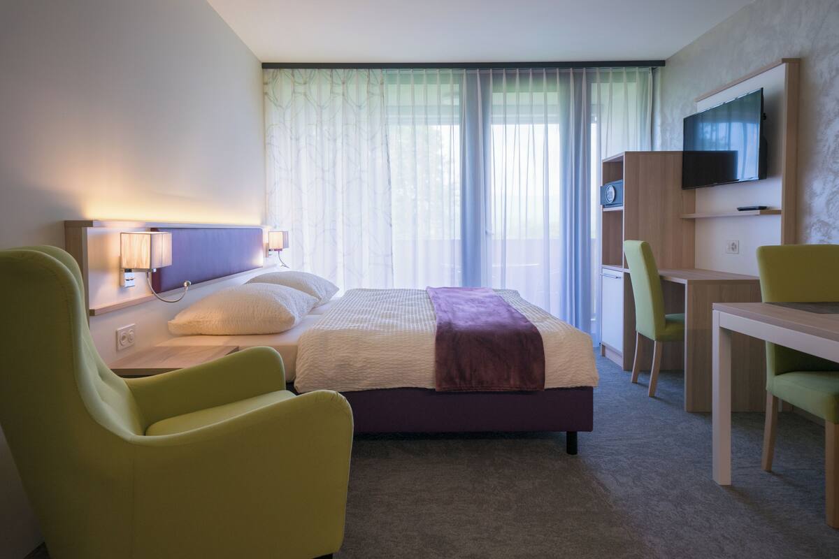 Ein Hotelzimmer mit einem Bett, zwei Stühlen, einem Schreibtisch, einem Fernseher und einem fenster mit Vorhängen.