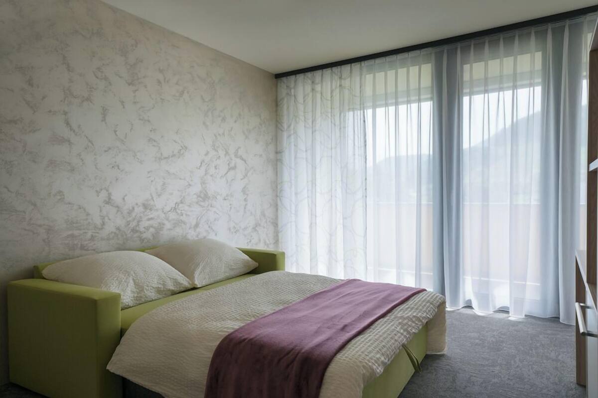 Ein Schlafzimmer mit einem Bett, einem grünen Sessel, einer Wand mit Marmoroptik und transparenten Vorhängen.