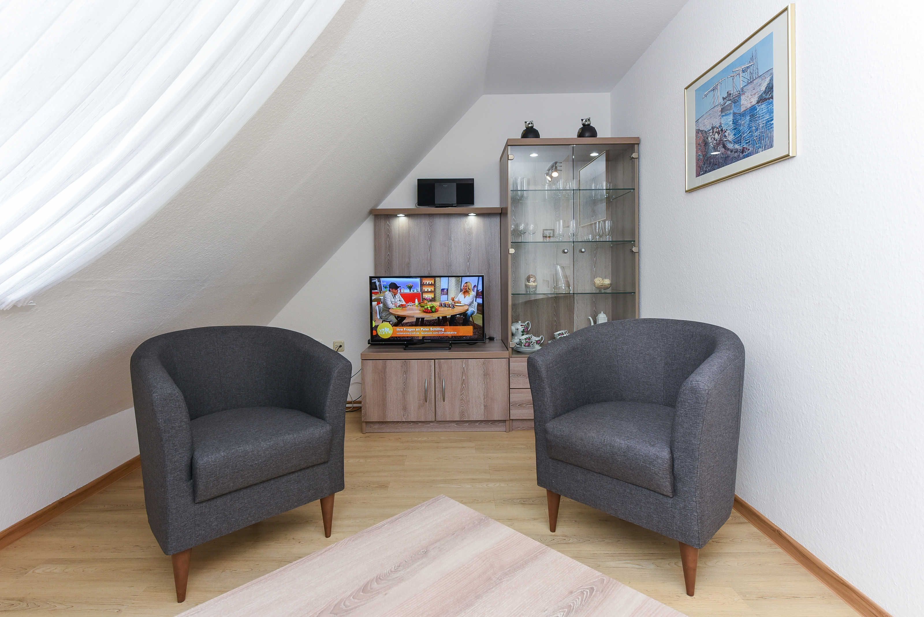 Moderni olohuone, jossa on kaksi harmaata nojatuolia, lasiovilla varustettu puinen kaappi, litteänäyttöinen televisio ja taulu seinällä.