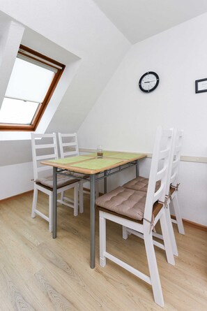 Ein kleiner, gut beleuchteter Raum mit schrägem Dach, einem Holztisch, weißen Stühlen, einer Uhr an der Wand und einem Fenster, das für natürliches Licht sorgt.