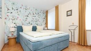 Ein Schlafzimmer mit Bett, Blumenwallpaper, einem Nachttisch, einer Lampe und einem Fenster mit Vorhängen.