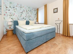 A bedroom with a bed, floral wallpaper, a side table, a lamp, and a window with curtains.