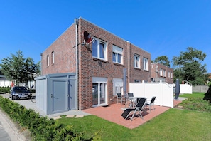 A two-story brick house with a patio area featuring a table and chairs, a car parked outside, and a small shed.