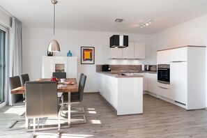 Une cuisine moderne avec des meubles blancs, un îlot central et des appareils électroménagers intégrés. On y trouve également un coin repas avec une table et des chaises en bois.