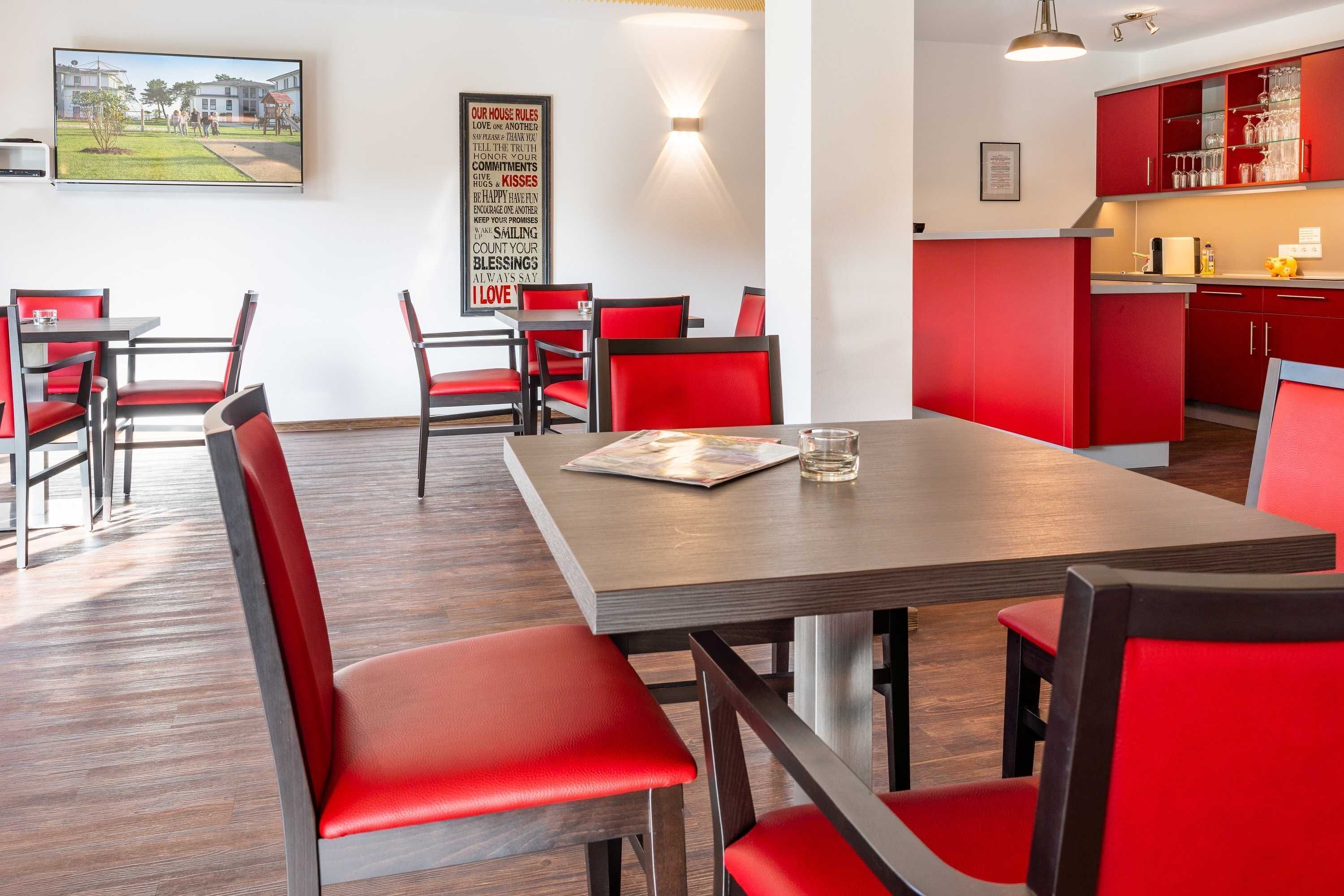 Een eetruimte met rode stoelen en een houten tafel, een keuken met rode kasten, en een tv aan de muur die een buitenscène toont.