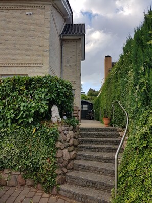 A residential building with a brick facade, a stone wall with greenery, and a staircase leading to a door.