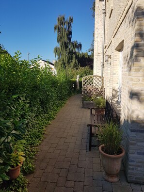 Ein gepflasterter Weg, gesäumt von üppiger Bepflanzung, einer hölzernen Bank und einem Backsteinhaus mit Gittertor.