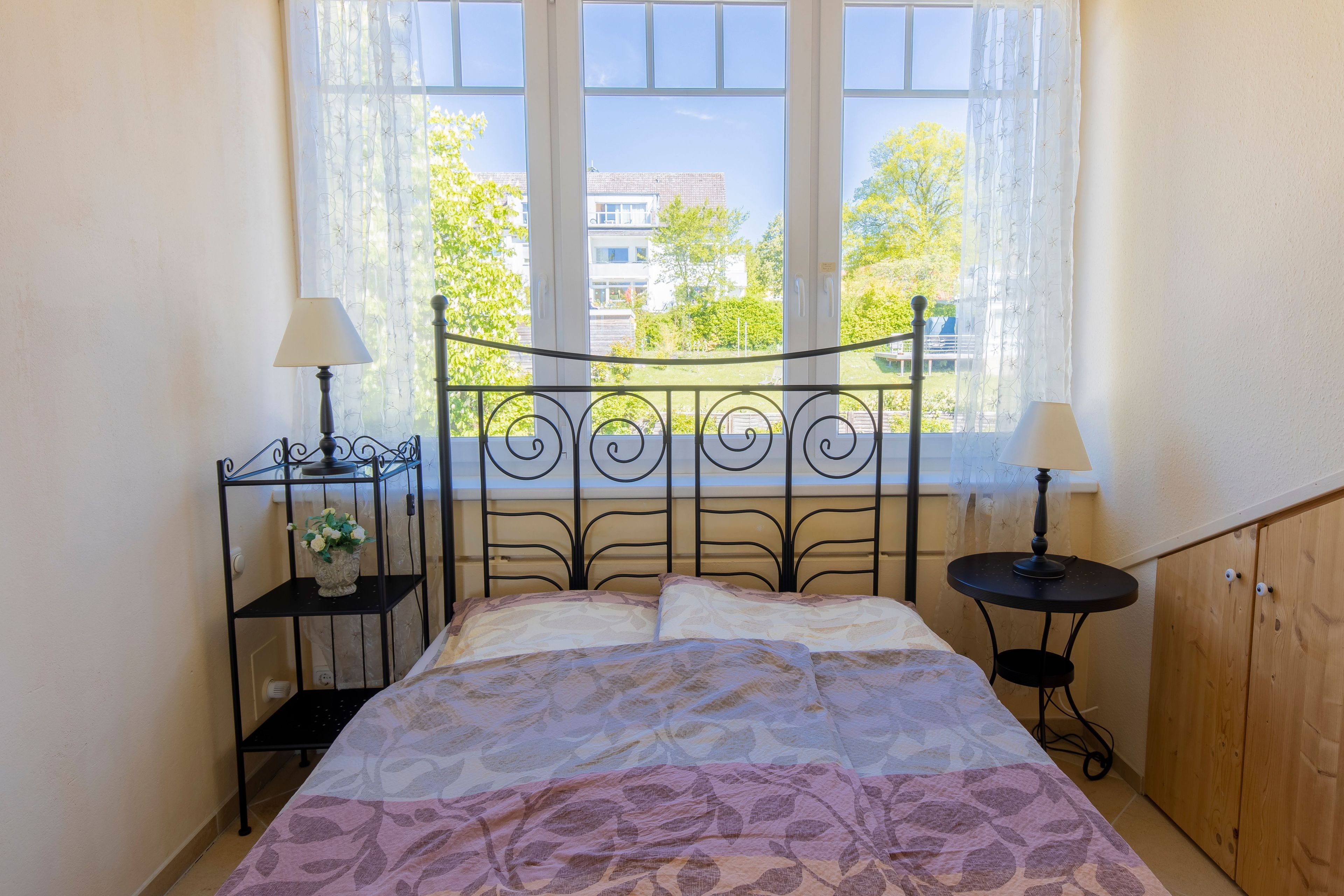 A bed with a patterned bedspread, two bedside tables with lamps, a small plant, and a window with a view of buildings and trees.