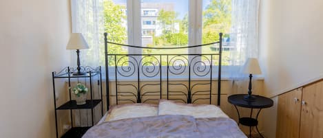 A bed with a patterned bedspread, two bedside tables with lamps, a small plant, and a window with a view of buildings and trees.