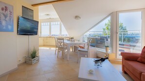 A modern living room with a dining area, a TV mounted on the wall, and large windows offering a view of the city. - Lighthouse - House Sun & Sea (Sierksdorf)