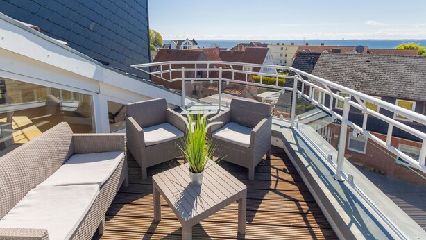 A deck with wicker furniture and a potted plant, overlooking a residential area and the sea. - Lighthouse - House Sun & Sea (Sierksdorf)