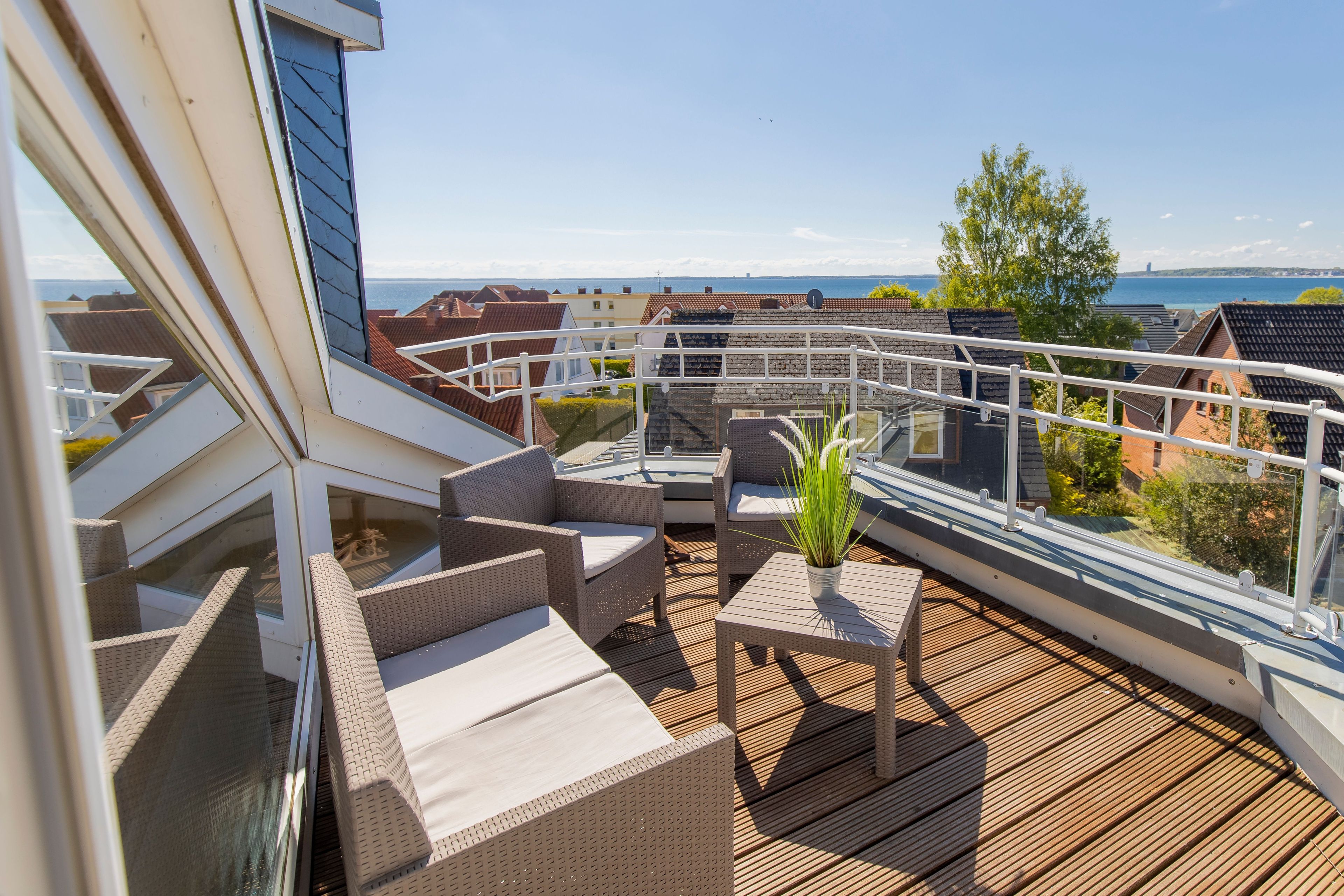 Een terras met rieten meubilair en een potplant, met uitzicht op een woonwijk en de zee.