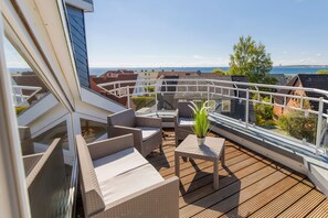 A deck with wicker furniture and a potted plant, overlooking a residential area and the sea.