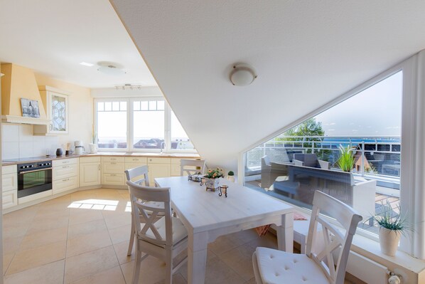 A modern kitchen with white cabinetry, a dining area with white chairs, and a large window overlooking a balcony with outdoor furniture.