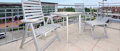A balcony with white metal chairs and a table, overlooking a parking area and buildings.