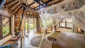 A room with a bed, a small table, and a chair. There is a window with a view of greenery outside. - Le Bamboo Bali (Pecatu)