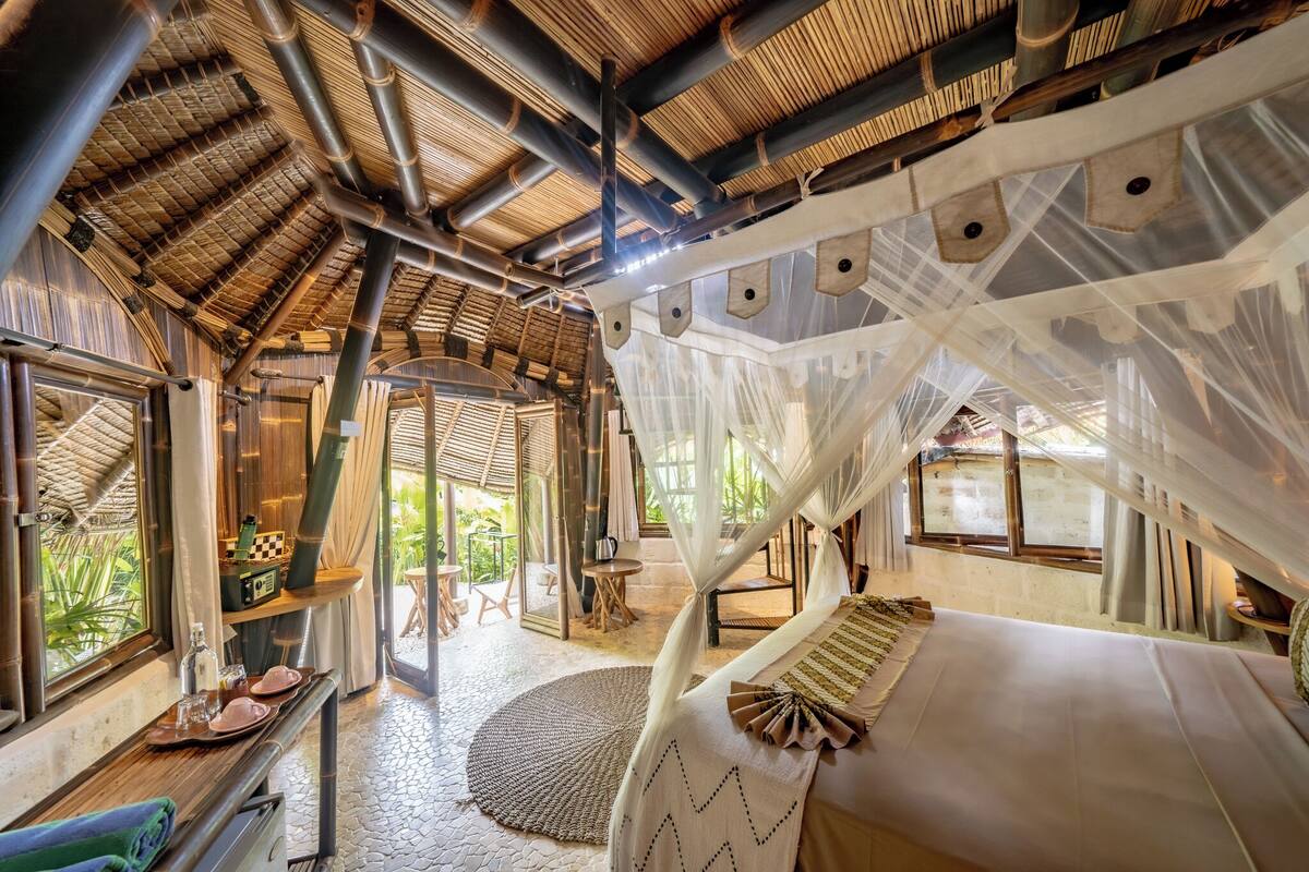 A room with a bed, a small table, and a chair. There is a window with a view of greenery outside.