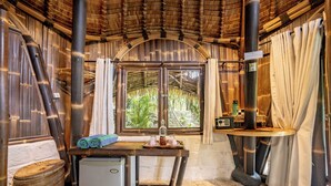 A rustic room with a thatched roof, a small table with a bottle and glasses, a window with curtains, and a woven basket. - Le Bamboo Bali (Pecatu)
