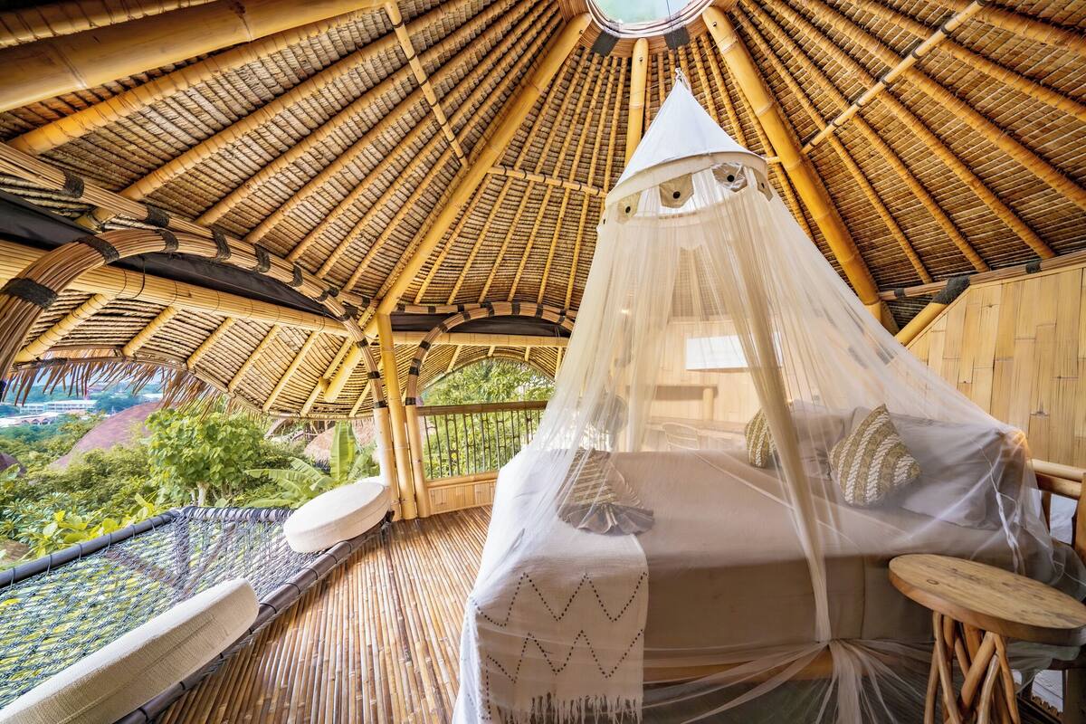 A thatched-roof room with a bed covered by a mosquito net, a small table, and a view of the surrounding landscape.