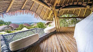 A thatched-roof outdoor area with bamboo flooring, cushioned seating, and a view of distant buildings. - Le Bamboo Bali (Pecatu)
