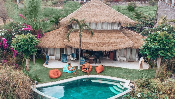 A thatched-roof house with a swimming pool, surrounded by lush greenery and colorful flowers. - Le Bamboo Bali (Pecatu)