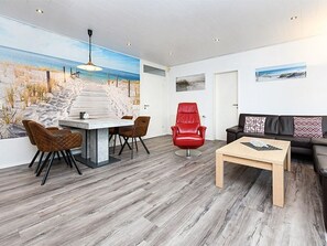 A modern living room with a wooden floor, a dining area with a white table and chairs, a red chair, a black sofa, and a painting of a beach scene on the wall.