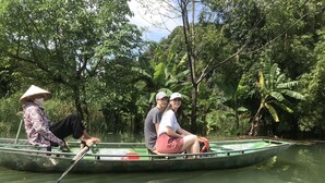 A narrow boat with three people, including a person in a conical hat, navigating through a waterway surrounded by dense greenery.