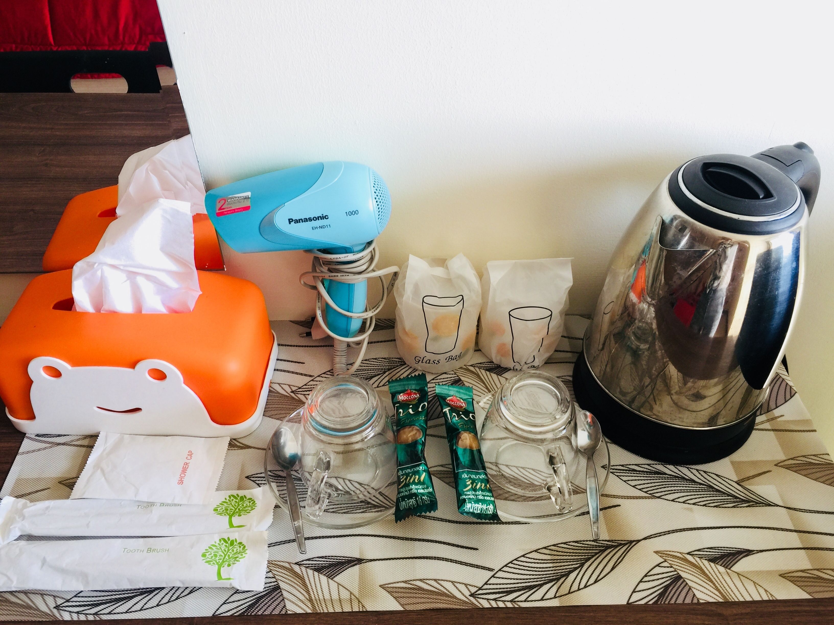 Disposición de baño con caja de pañuelos, portalápices para cepillos de dientes, tarro de cristal, cepillo de dientes, taza, cuchara, sobre de té, vaso y hervidor.