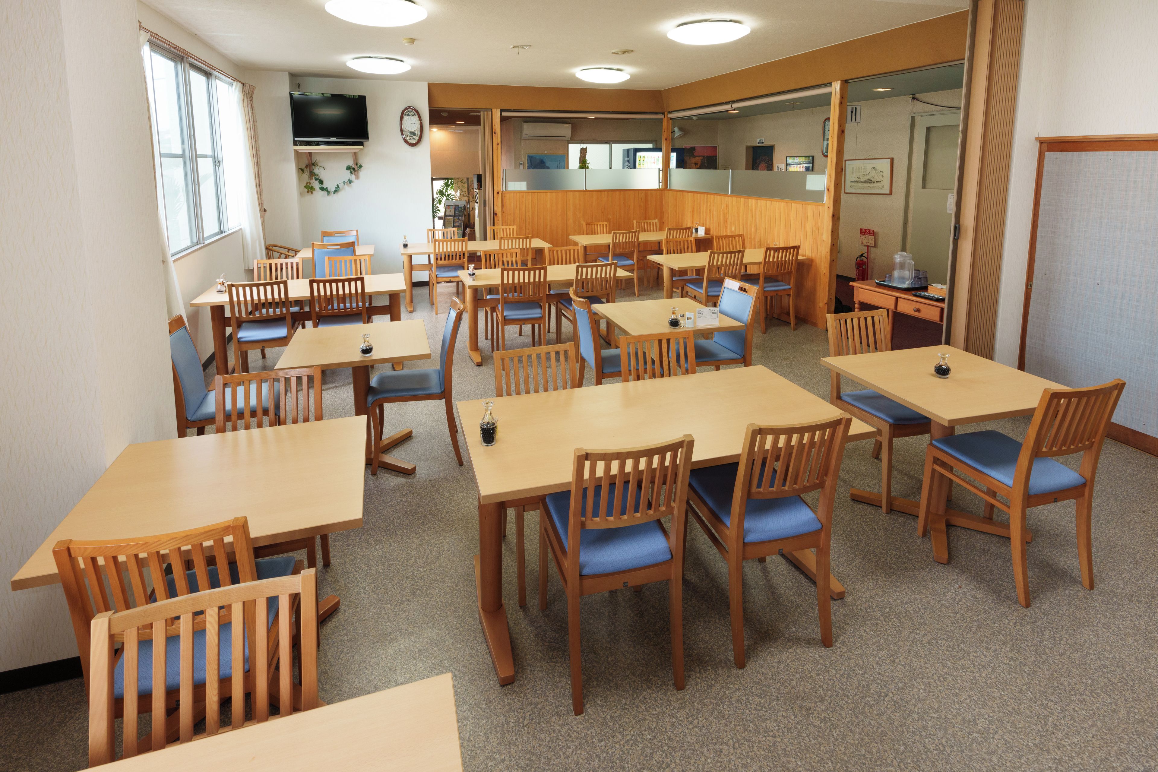 A dining area with multiple wooden tables and chairs, a television on the wall, and a view of the outside through a window.