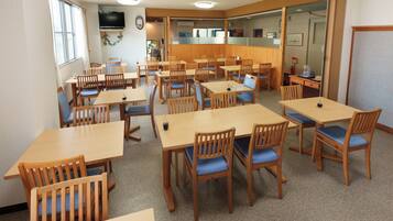 A dining area with multiple wooden tables and chairs, a television on the wall, and a view of the outside through a window.