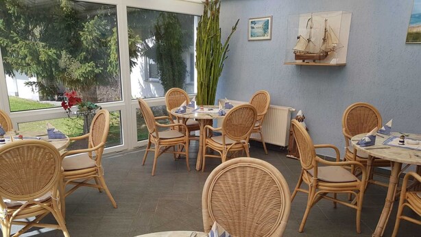 A dining area with wicker chairs and round tables, large windows with a view of greenery, and nautical-themed decor on the walls. - Small Bungalow Wiesenperle - Small Bungalow meadow pearl (Trassenheide)